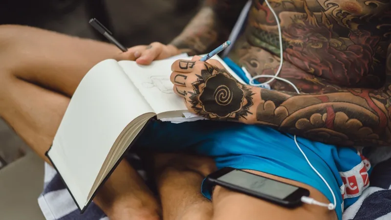 A heavily tattooed person sketches in a notebook with one hand, a phone resting on a striped towel in their lap and white earbud cables trailing up to their over-ear headphones.