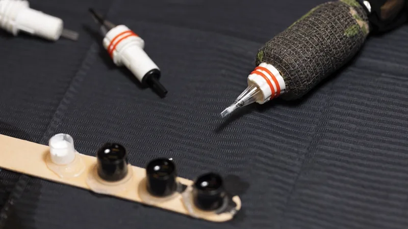 Three tattoo machine cartridge needles and a short row of small black ink caps on a dark fabric mat. One cartridge is wrapped in dark green grip tape.